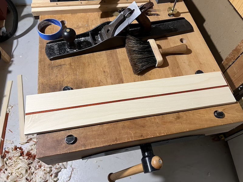 A workbench with a number 7 hand plane, brush, and jointed board of poplar with a thin stripe of padauk down the center.