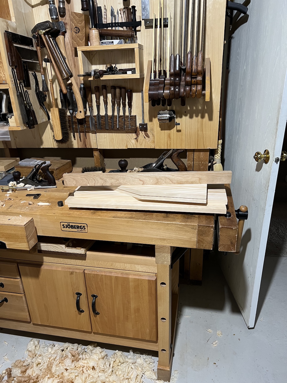 A stack of cut wood pieces laying on a workbench, with a tool wall covered in chisels, saws, and planes in the background.