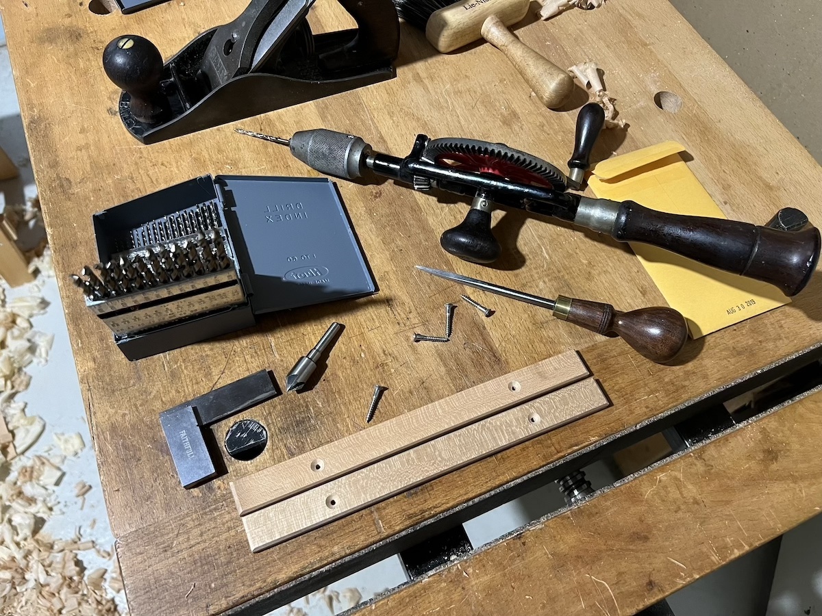 Two long and thin pieces of wood with countersunk screw holes at either end resting on a workbench amonstly scattered screws, an awl, hand plane, and hand drill.