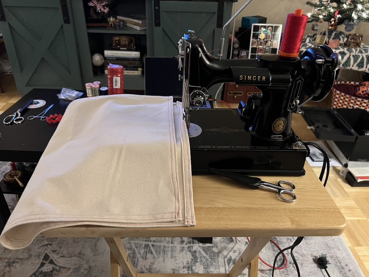 A portable Singer 221K sewing machine with a shiny black finish and gold decals sits on a small folding table with a piece of natrual canvas and ends hemmed in red thread.